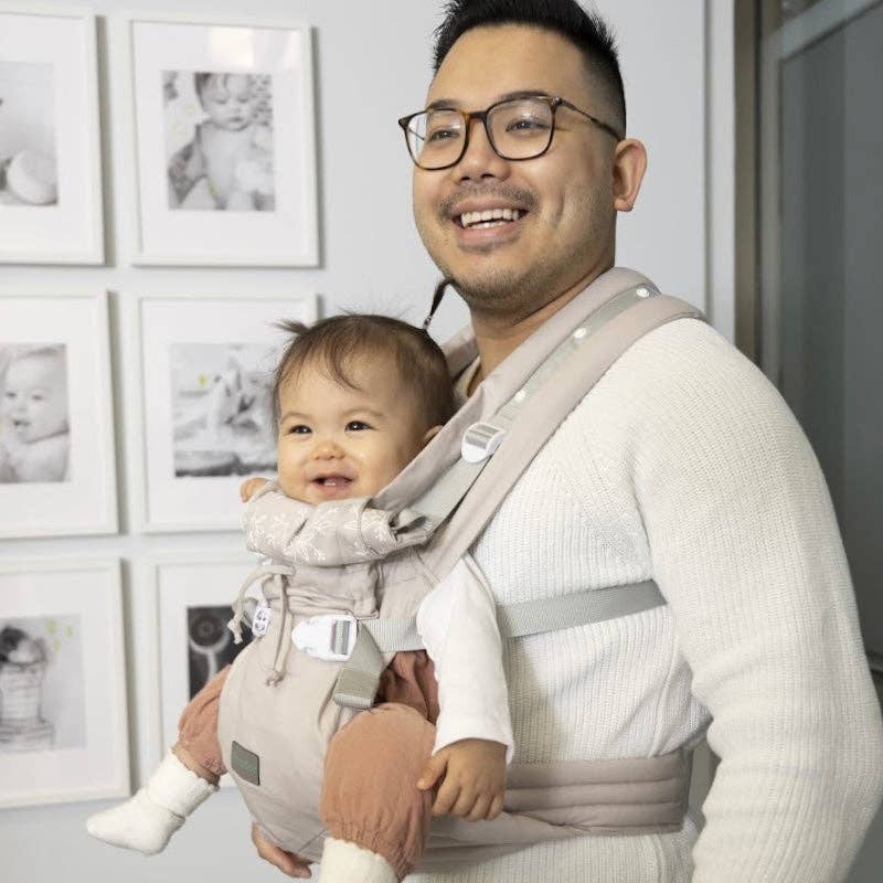 Man holding a baby in a carrier with framed pictures on the wall.