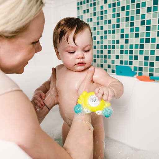 Baby in a bath with a toy, surrounded by a tiled bathroom.