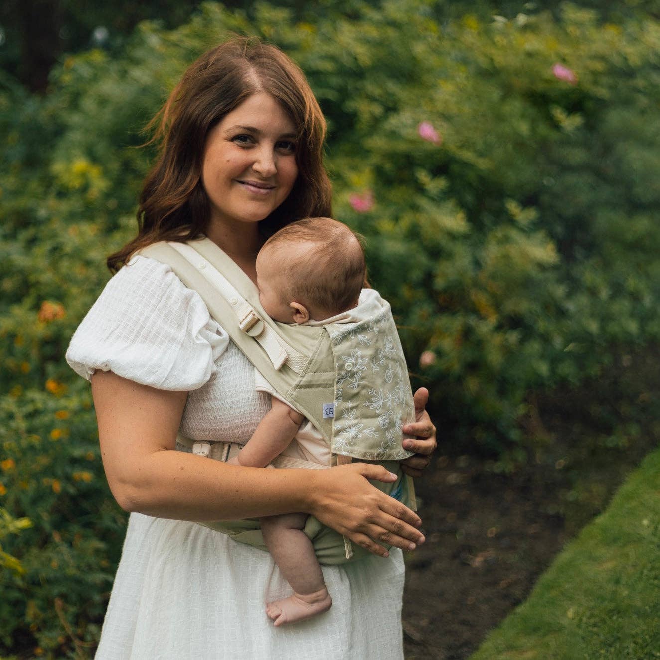 Woman holding a baby in a sling outdoors with greenery in the background