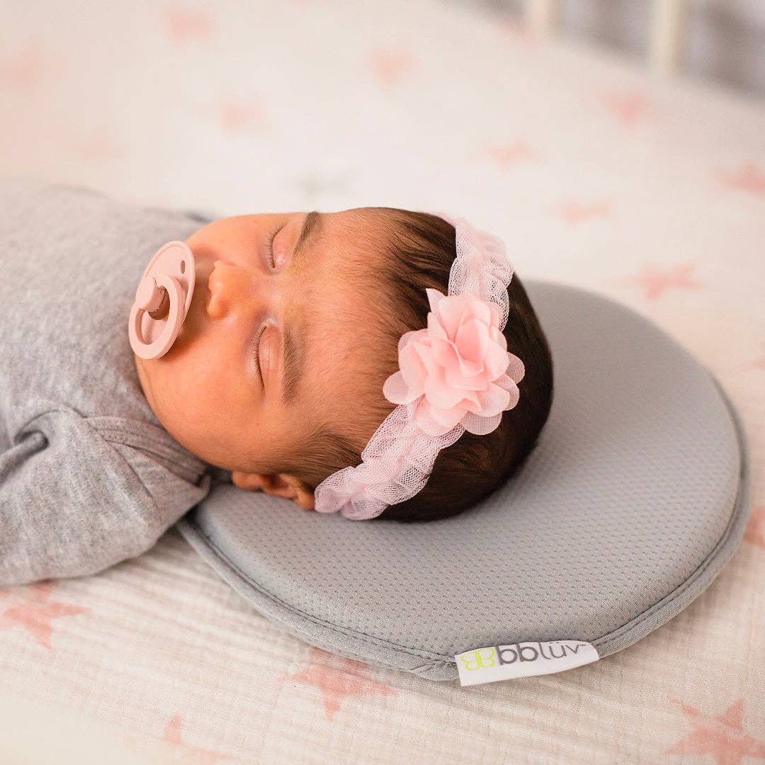 Newborn baby sleeping on a gray pillow with a pink flower headband, brand 'bbjluv' visible.