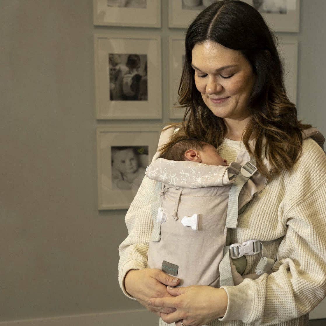 Woman holding a baby in a carrier with framed photos on the wall.