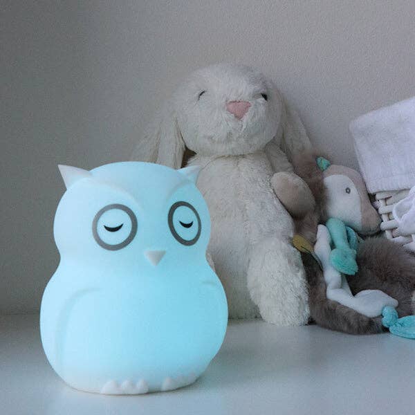 Owl-shaped night light emitting a soft blue glow, placed on a surface with plush toys in the background.