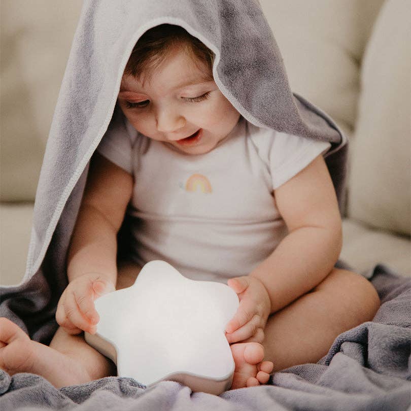 Baby sitting on a couch under a gray blanket holding a white star-shaped night light.