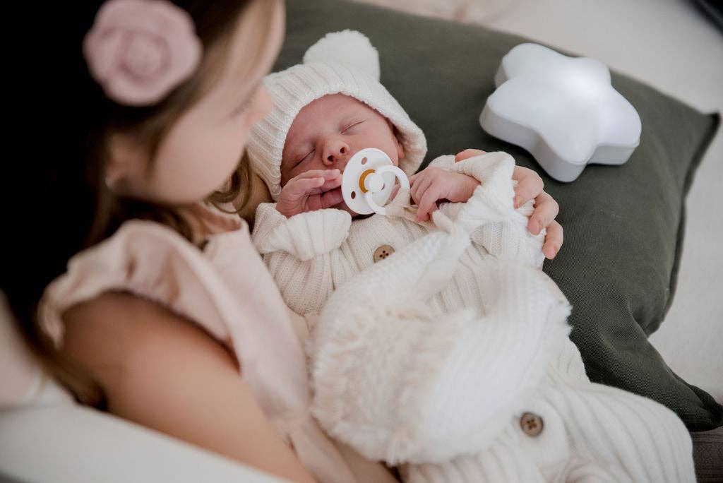 Newborn baby in a white outfit with a pacifier, held by an adult, with a soft light in the background.