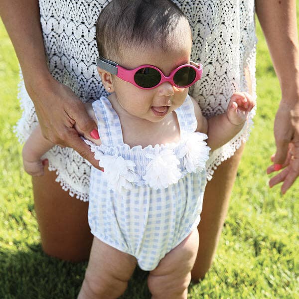 Baby in a checkered romper and pink sunglasses standing on grass with a person holding their hand.