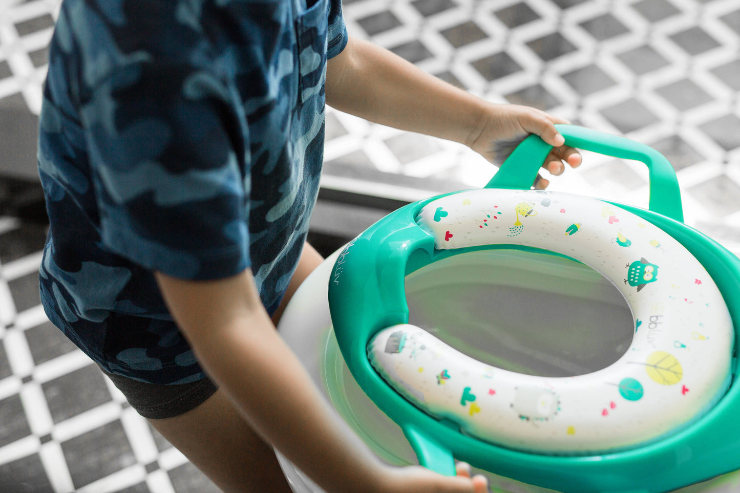 Child holding a green and white potty seat with colorful patterns on a tiled floor.