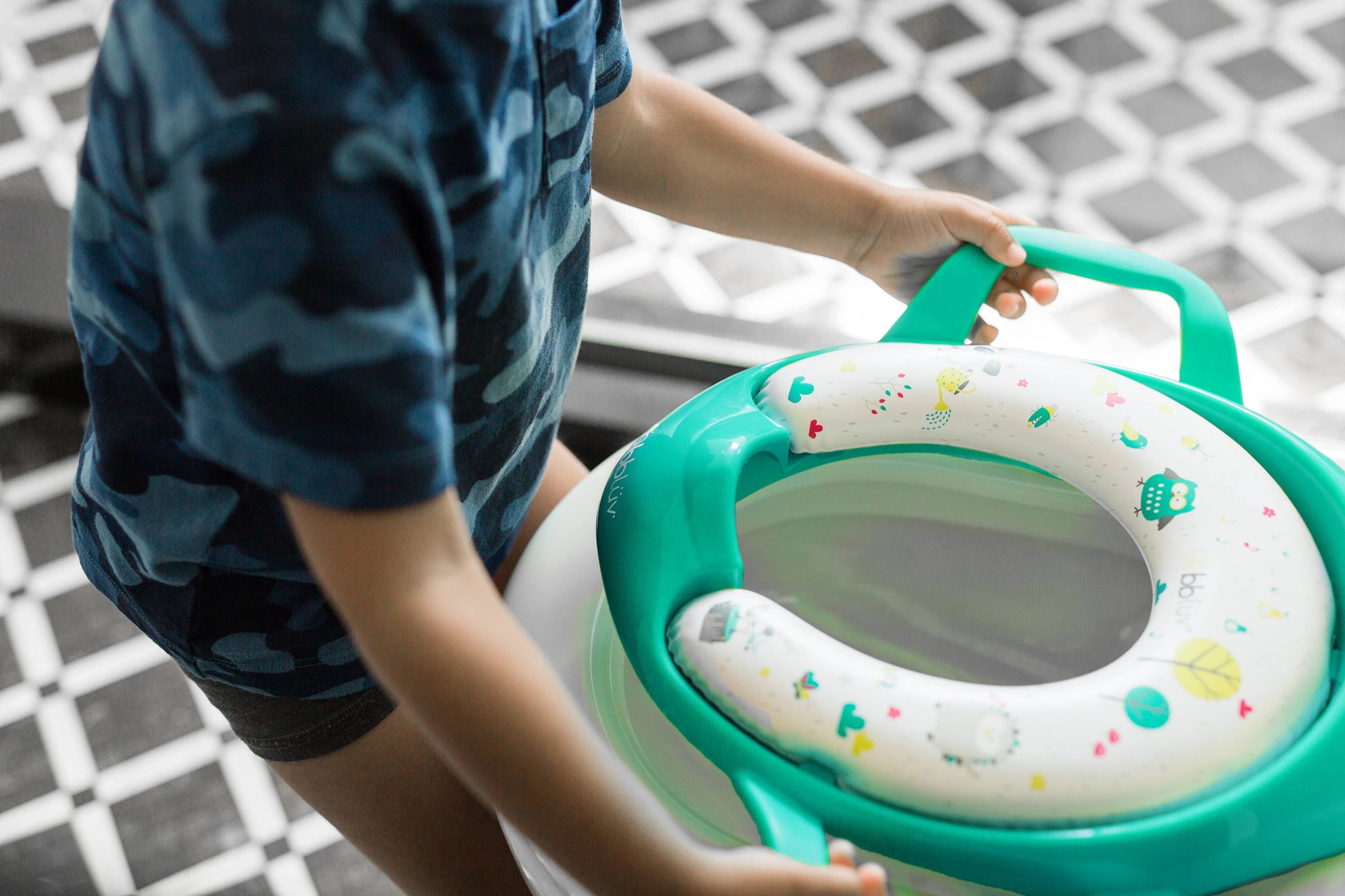 Child holding a green and white potty seat with colorful patterns on a tiled floor.