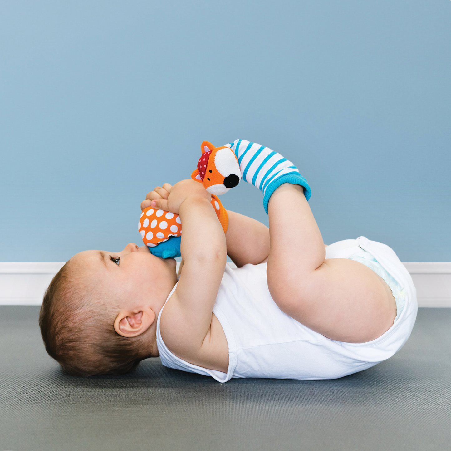 Baby lying on a gray floor with a toy and colorful socks against a light blue wall.