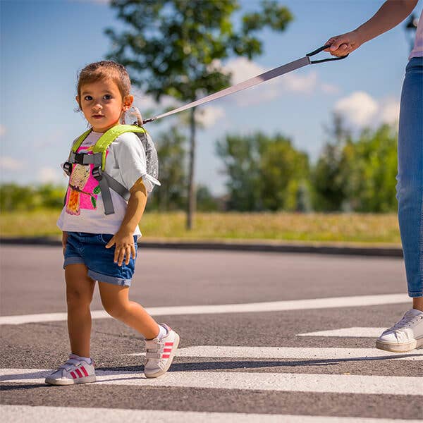 Child wearing a backpack and being led by a leash on a street with trees in the background