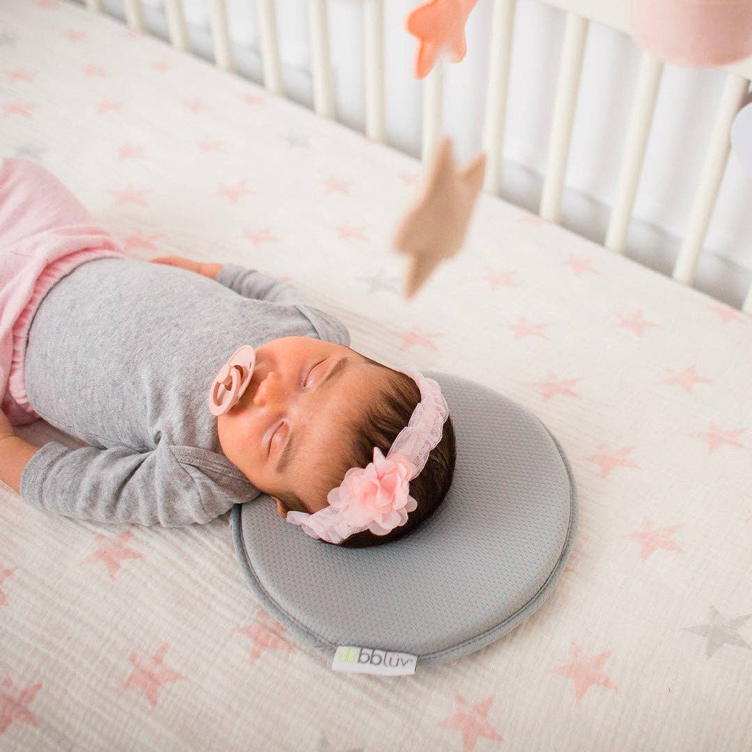 Newborn baby lying on a gray head support pillow with pink floral headband in a crib.